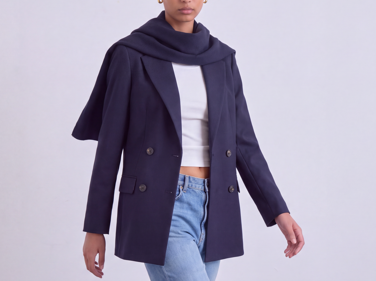 A model wearing a navy blue double-breasted coat with a draped collar, styled over a white top and light blue jeans, walking against a light gray studio background.